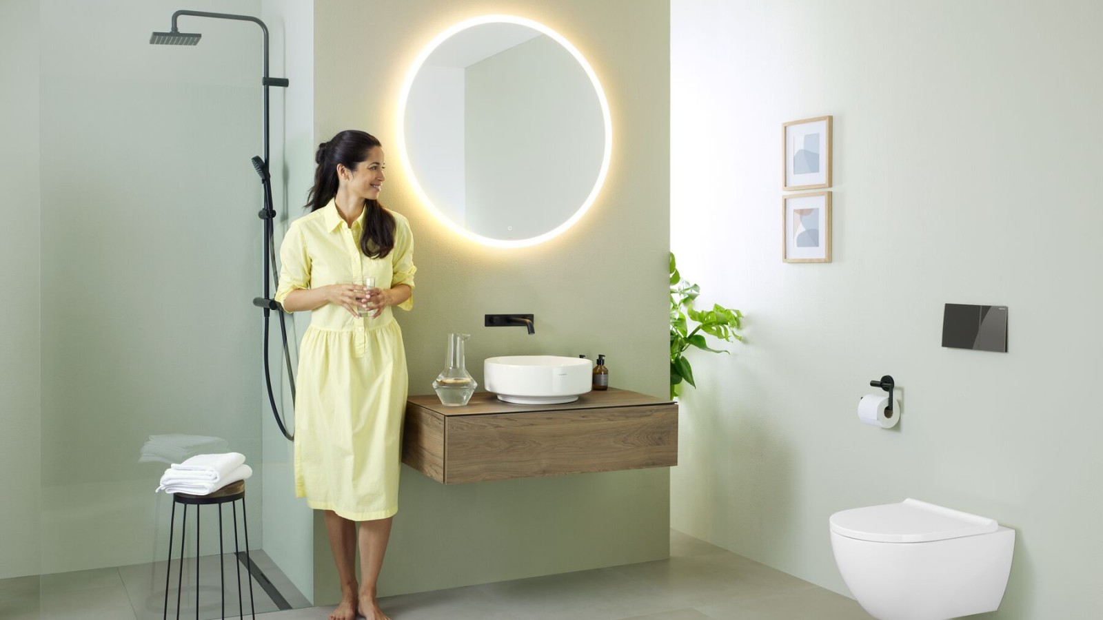 Mujer con vestido amarillo de pie delante de un baño verde menta con muebles y cerámica de baño de Geberit y grifería negra. Mujer con vestido amarillo de pie delante de un baño verde menta con muebles y cerámica de baño de Geberit y grifería negra.
