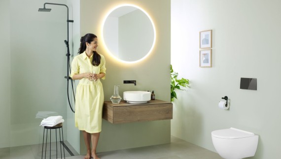 Mujer con vestido amarillo de pie delante de un baño verde menta con muebles y cerámica de baño de Geberit y grifería negra.