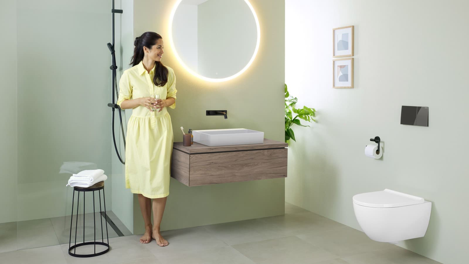 Mujer con vestido amarillo de pie delante de un baño verde menta con muebles y cerámica de baño de Geberit y grifería negra. Mujer con vestido amarillo de pie delante de un baño verde menta con muebles y cerámica de baño de Geberit y grifería negra.